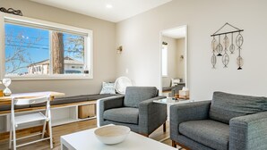 A modern living room with a large window, grey sofas, a white table, and a decorative wall hanging.