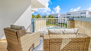 A balcony with wicker furniture, a pool, and a view of modern buildings. - Steps from a beautiful beach – Your Puerto Rican Getaway Awaits (Río Grande)