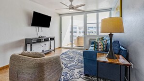 A modern living room with a flat-screen TV, a ceiling fan, a sofa, a coffee table, and a large window offering a city view. - Steps from a beautiful beach – Your Puerto Rican Getaway Awaits (Río Grande)