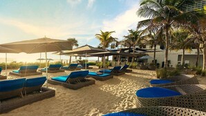 A beachside lounge area with sun loungers and umbrellas, palm trees, and a building in the background. - Luxury Waterfront Condo (Hallandale Beach)