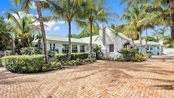 A white building with a green roof, surrounded by palm trees and a brick pathway.