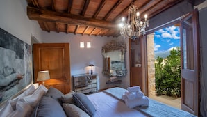 A bedroom with a large bed, a chandelier, a mirror, and a view of greenery outside.
