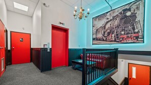 Eine Hotellobby mit einem roten Aufzug, einer blauen Wand mit einem Zuggemälde, einer schwarz-roten Bank und einem Kronleuchter.