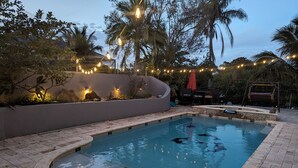 Une piscine à bord incurvé, entourée d’une terrasse en briques. On aperçoit des guirlandes lumineuses et des palmiers en arrière-plan.