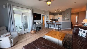 A modern living room with a sofa, a brown leather ottoman, a kitchen area with a bar, and a ceiling fan. - *New*Oceanview, Golf, Pool, Tennis, Beach Access (Corolla)