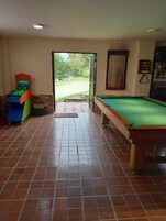 Una habitación con mesa de billar, mesa de snooker y dardos. Hay una puerta que da a un área exterior con vista a un campo de golf.