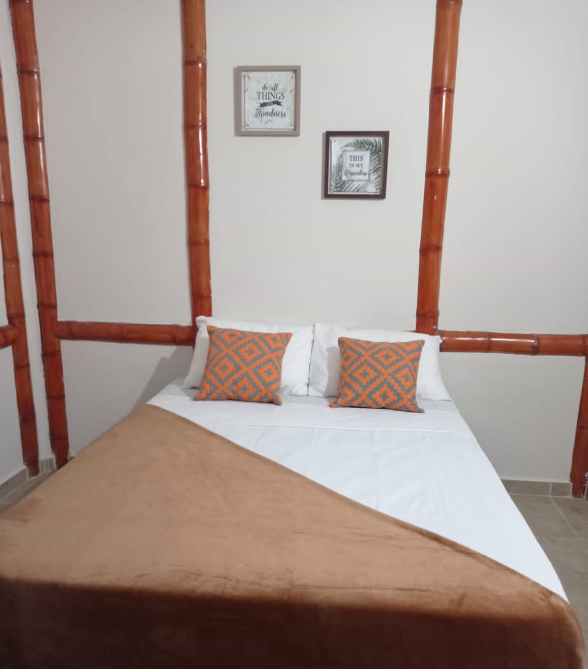 A bed with a white mattress and two orange patterned pillows, a brown blanket, and bamboo headboard railings.