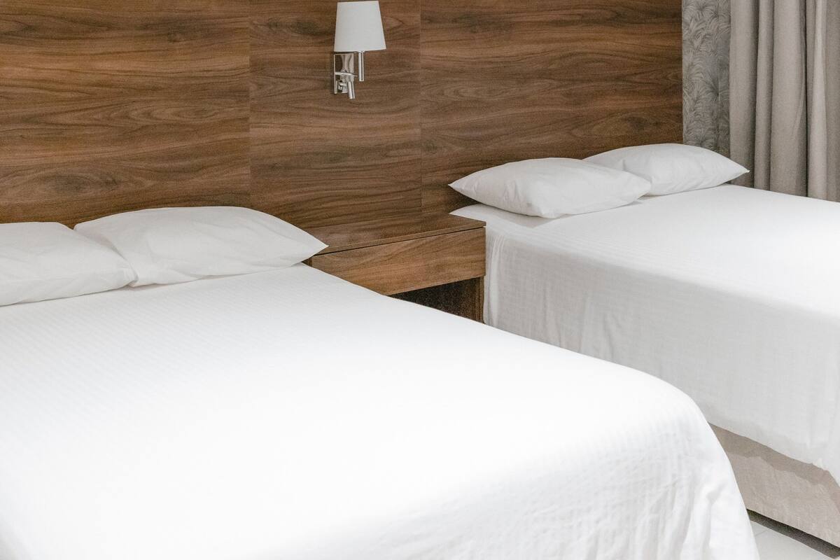 Habitación de hotel con dos camas, cabecero de madera, lámpara de pared y papel tapiz estampado.