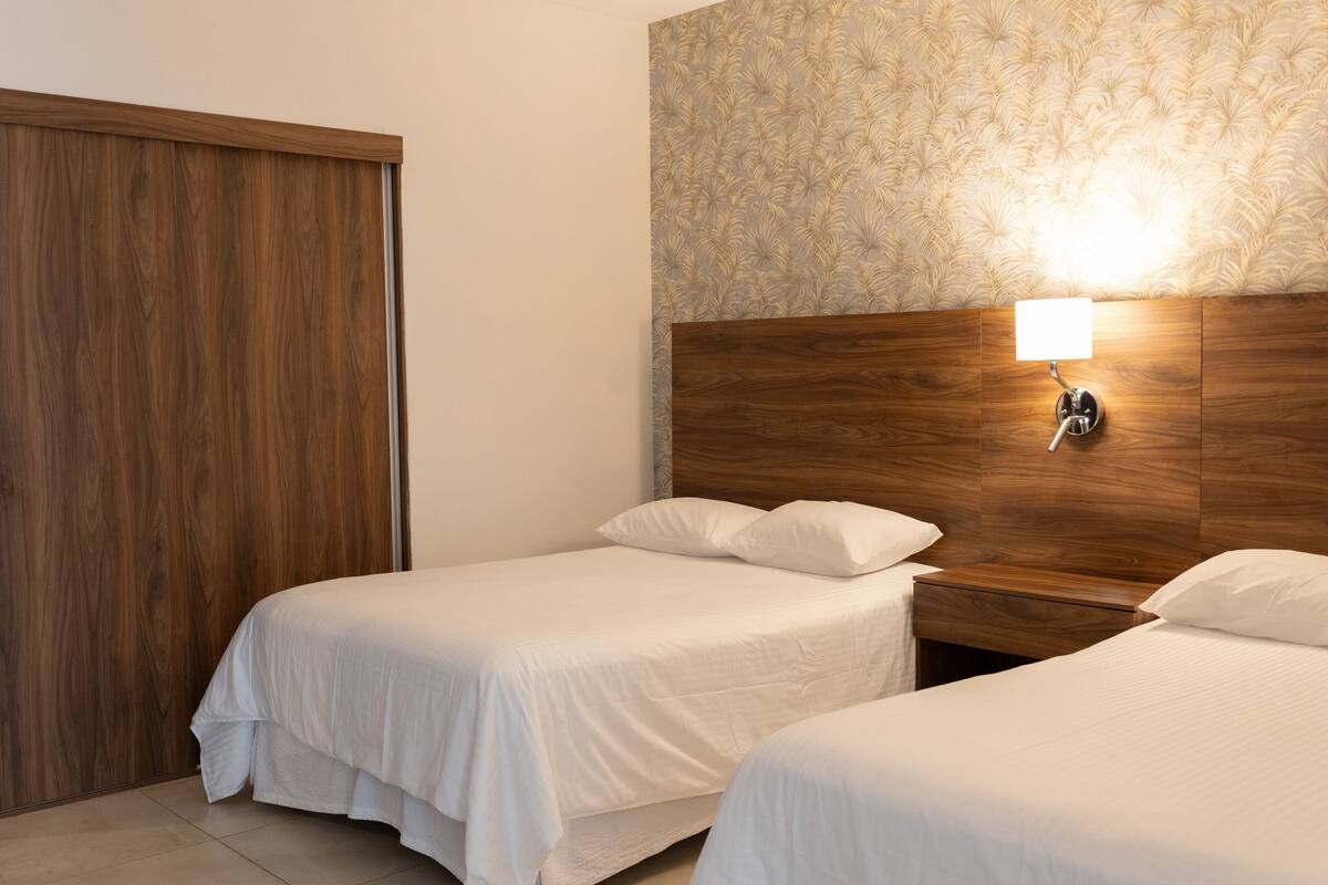 Una habitación de hotel con dos camas individuales, cabeceros de madera, una mesita de noche con lámpara y papel tapiz estampado en la pared.