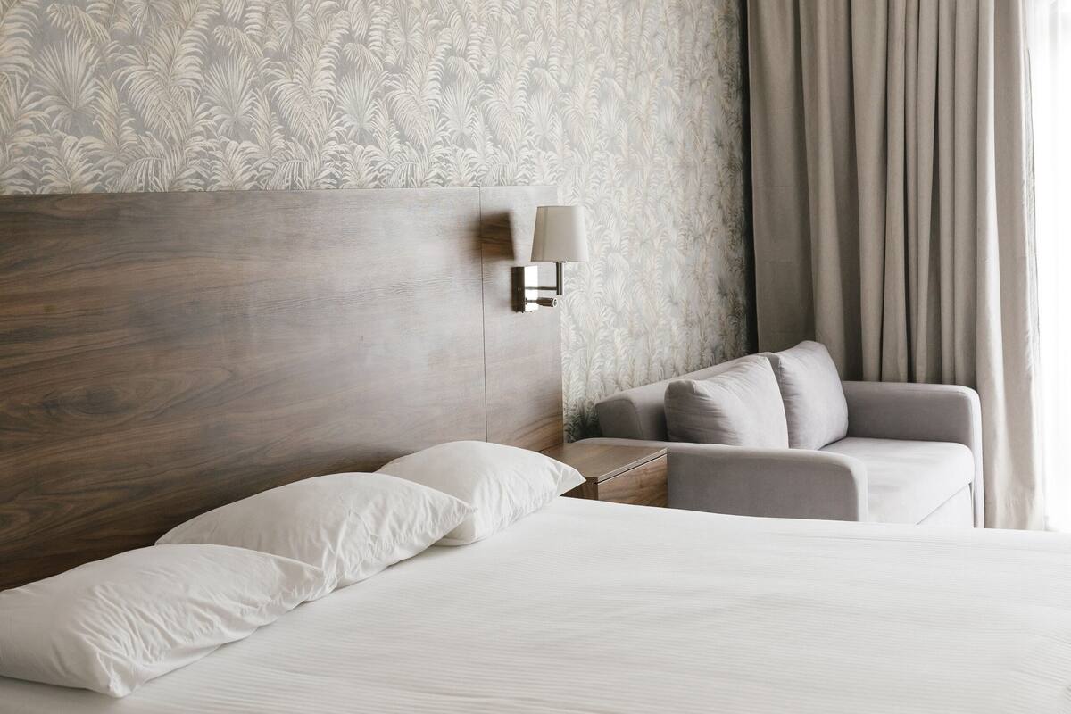 Una habitación de hotel con una cama grande, una zona de estar con un sillón, una lámpara de pared y una ventana con cortinas.