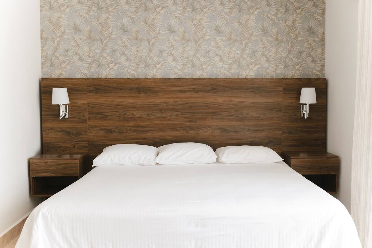 Una cama bien tendida con sábanas blancas, un cabecero de madera y dos mesitas de noche con lámparas.