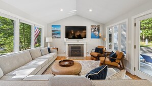A spacious living room with a large sofa, a coffee table, and a fireplace. There are multiple windows providing natural light and views of the outdoors.