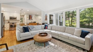 A modern living room with a sectional sofa, a coffee table, and a view of the kitchen and outdoor area.