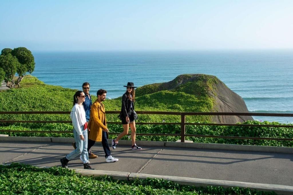 Un sendero pavimentado con barandilla, con vistas a un paisaje costero con colinas verdes y el mar.