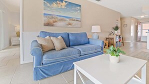 A living room with a blue sofa, a white coffee table, and a painting of a beach scene on the wall. - Beach Condo |  Pool | 2 Bedrooms | Monthly Rental (Cape Canaveral)