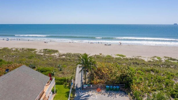 A beach view with a clear sky, a sandy beach, and a few people. There is a building with a roof and a pathway leading to the beach. - Beach Condo |  Pool | 2 Bedrooms | Monthly Rental (Cape Canaveral)
