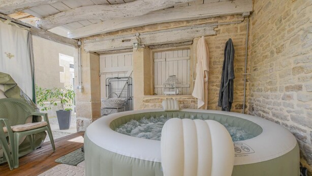 A rustic room with a stone wall, a wooden ceiling, a green chair, a white bathtub, and a window with a view.