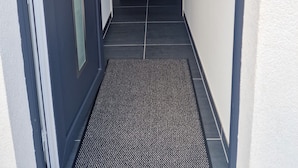 A narrow hallway with a dark door on the left, a mat at the entrance, and a kitchen area with a refrigerator in the distance. - Im Stadtzentrum, in der Nähe von Musikschulen (Trossingen)