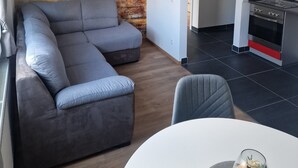 A modern living room with a grey sectional sofa, a white dining table with a glass centerpiece, and a kitchen area with a stainless steel oven. - Im Stadtzentrum, in der Nähe von Musikschulen (Trossingen)