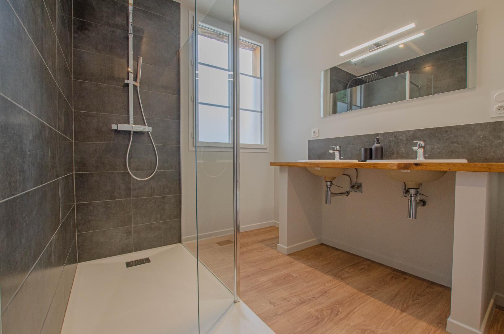 Een moderne badkamer met een glazen douchecabine, een houten wastafelmeubel met een wastafel en een grote spiegel erboven.