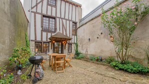 Un pati amb taula i cadires de fusta, una barbacoa i un jardí ple de flors i plantes.