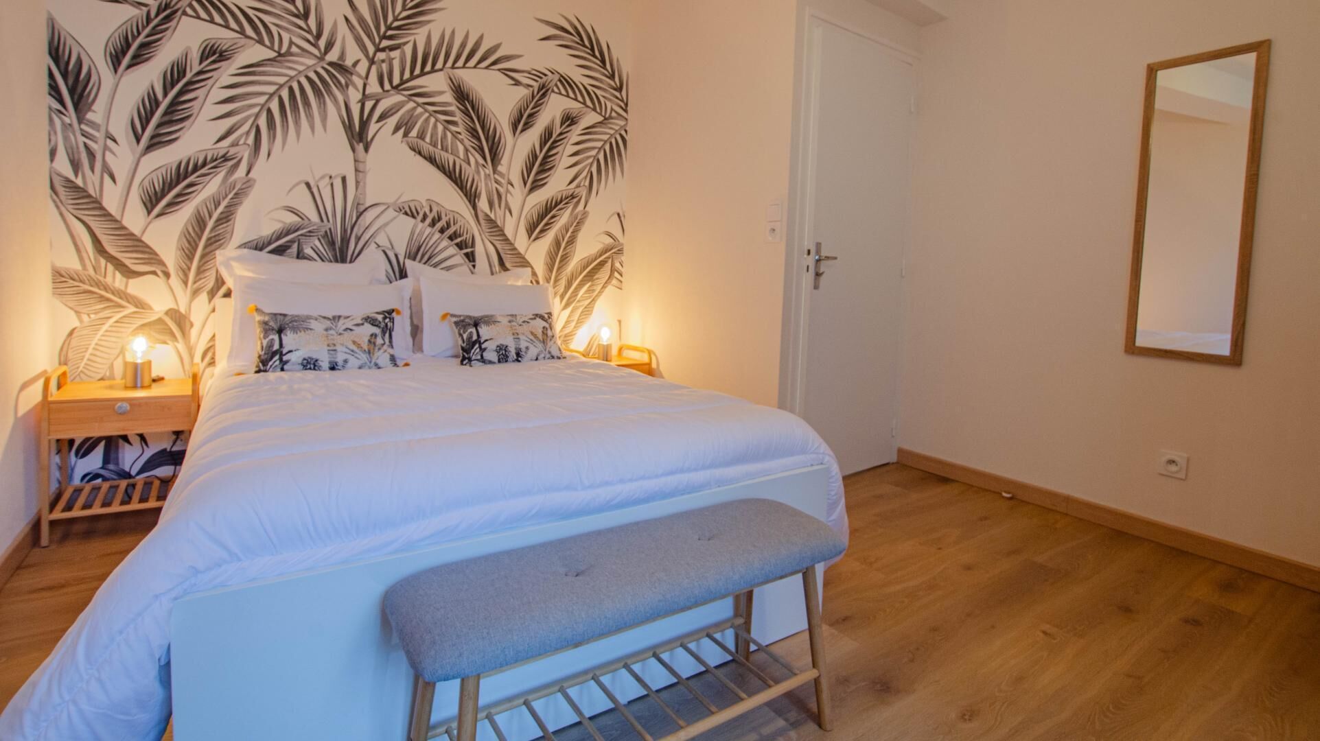 Quarto com cama, um banquinho, mesa de cabeceira, abajur e parede com padrão de folhagem.