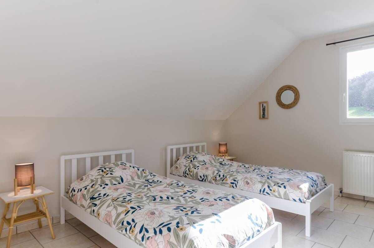 A bedroom with two single beds, a small table with a lamp, a window with a view, and a wall-mounted mirror.