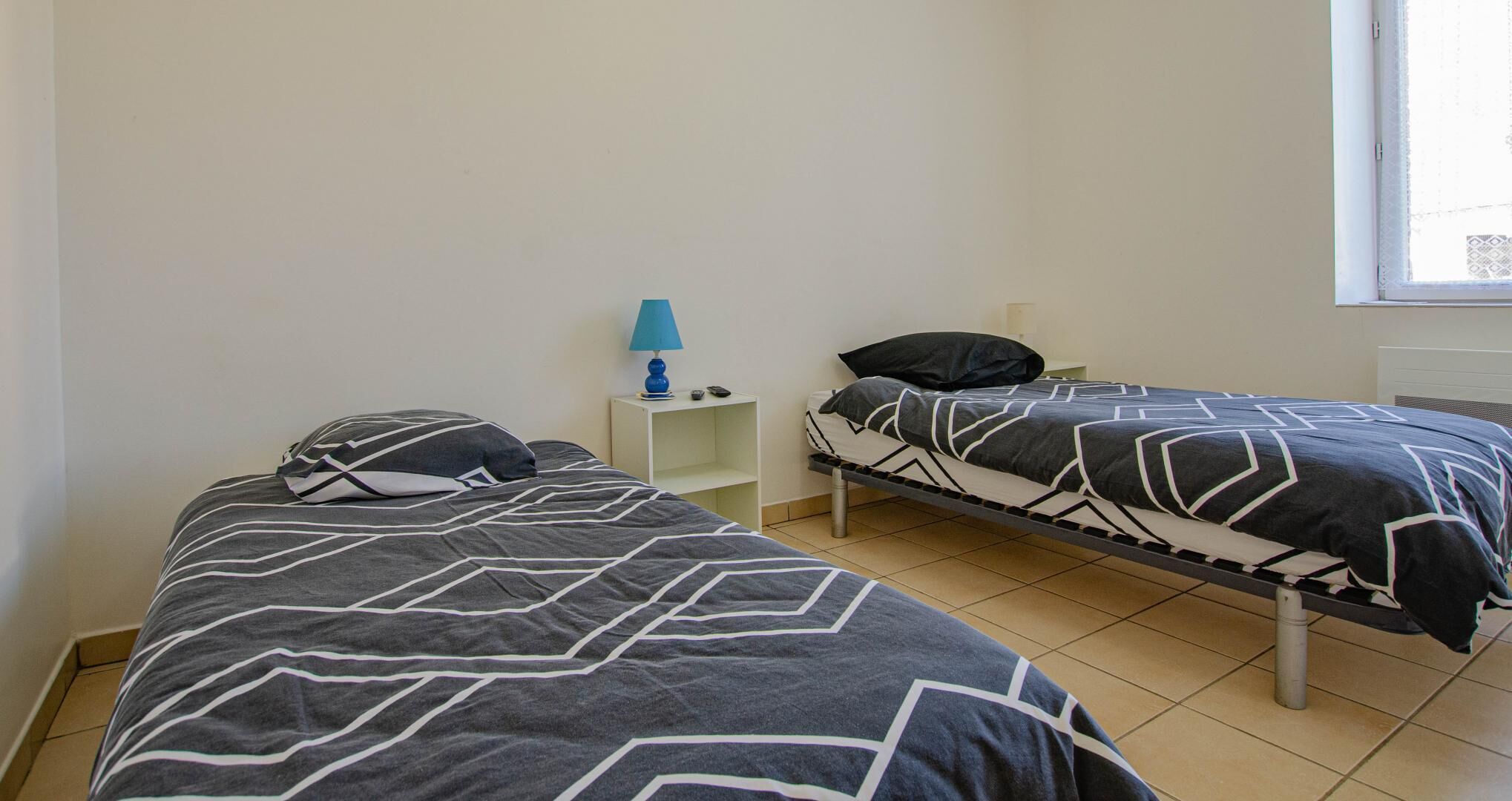 A small, clean room with two single beds, a nightstand with a lamp, and a window.