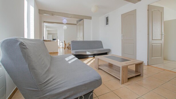 Un salon avec un canapé gris, une table basse en bois et un fauteuil gris. Il y a une porte sur la droite et une fenêtre sur la gauche.