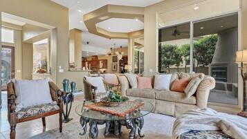 A spacious living room with a glass coffee table, a sofa, and a chair. There is a kitchen in the background with large windows.