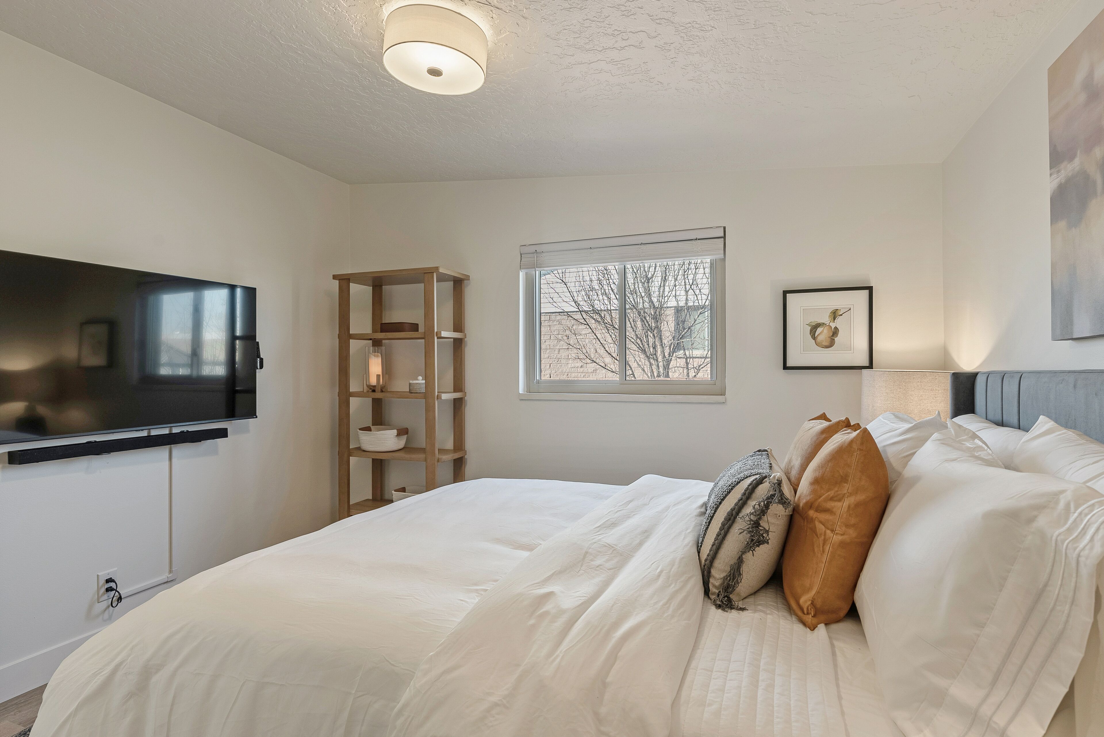 A bedroom with a bed, a TV mounted on the wall, a window with a view of trees, and a shelf with decorative items.