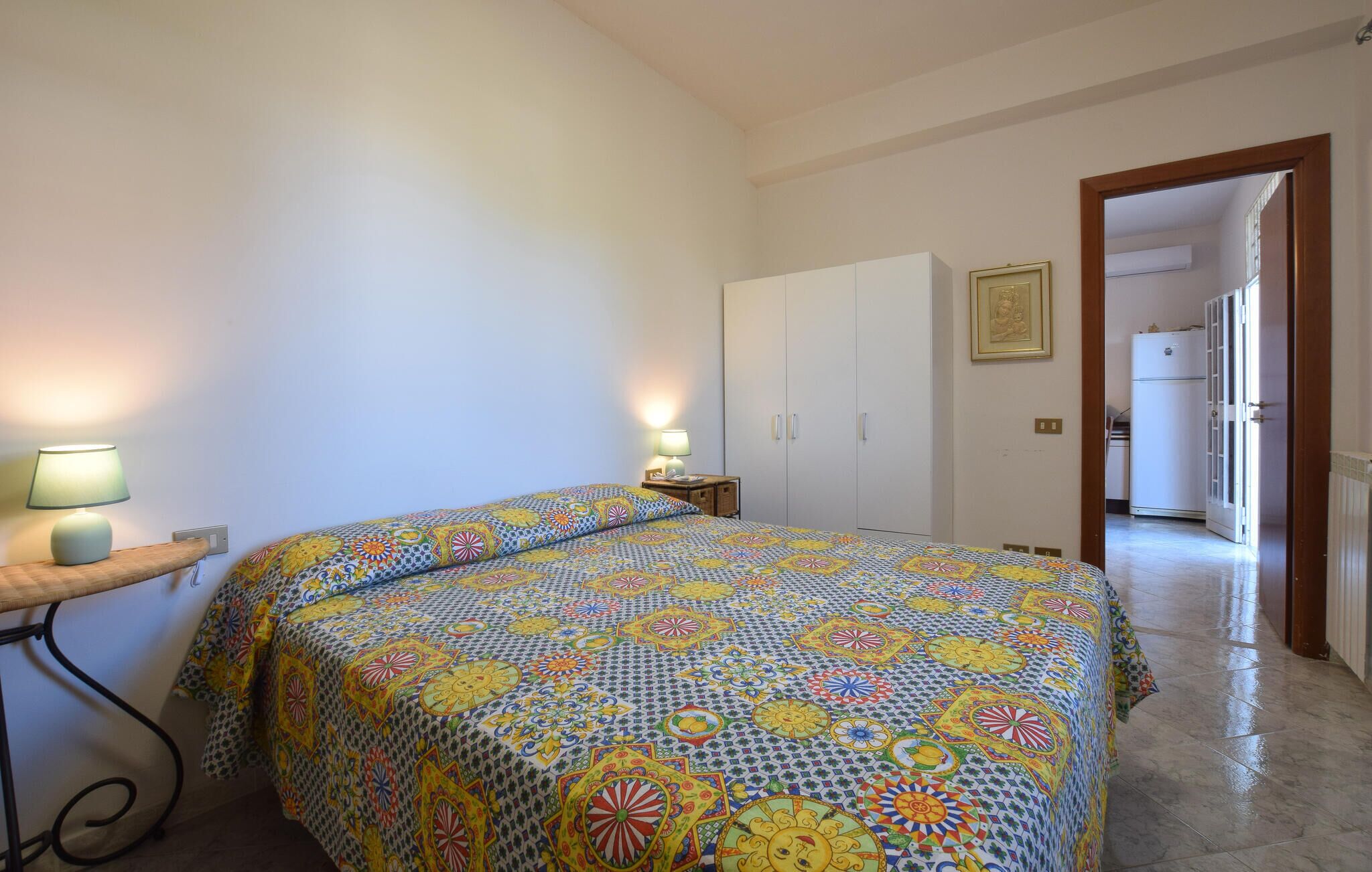 A bedroom with a bed, a nightstand, a wardrobe, and a door leading to a kitchen.