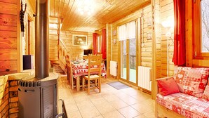 A wooden cabin interior with a central wood-burning stove, a dining area with a table and chairs, and a sofa with patterned cushions. - Le Chant de la Rivière - Chalet 3* pour 8 personnes (Ventron)