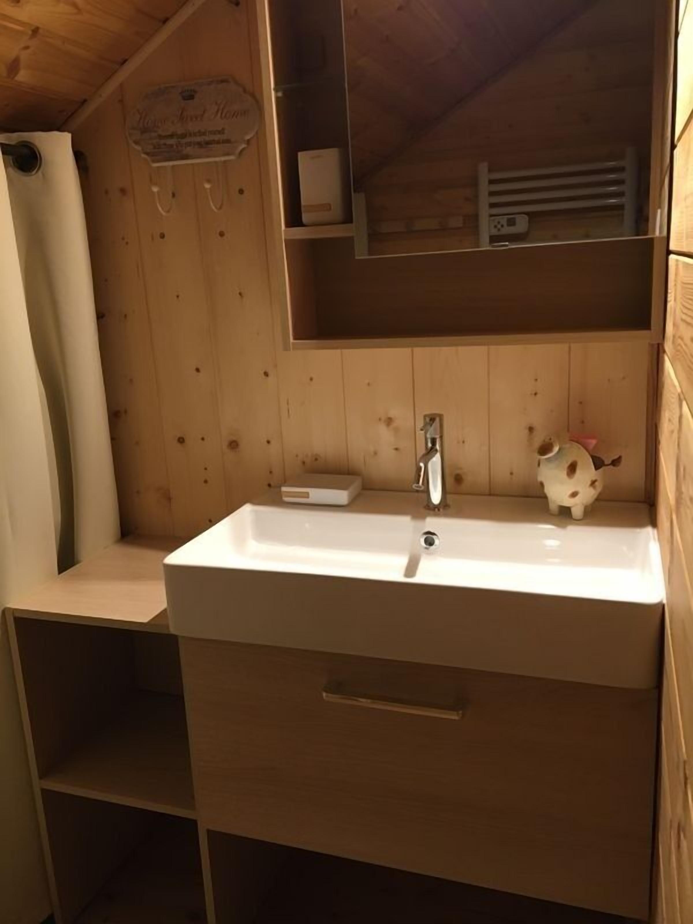 Bany d'una cabana de fusta amb un vàter blanc, un mirall i una petita estant amb un objecte decoratiu.