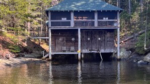 Eine Holzhütte mit grüner Dachdeckung, zwei Etagen und einem Steg, der in einen von Bäumen gesäumten Fluss führt.
