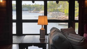 Ein Zimmer mit Holzboden, einer Couch mit gestreiften Kissen, einer Lampe und Blick auf einen See mit Steg und Bäumen.