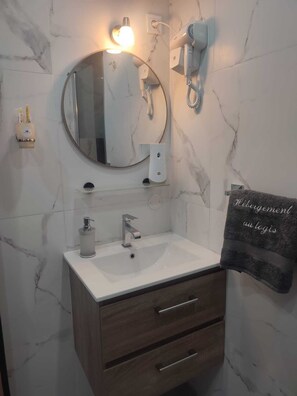 A bathroom with a marble wall, a round mirror, a sink with a modern faucet, a towel rack with a towel, and a hairdryer mounted on the wall. - Hébergement AU Logis (Sougé-le-Ganelon)