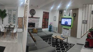Un salon moderne avec un canapé d’angle gris, un téléviseur à écran plat, une cheminée et un coin repas comprenant une table et des chaises.