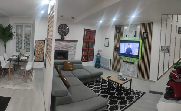 A modern living room with a grey sectional sofa, a flat-screen TV, a fireplace, and a dining area with a table and chairs. - Hébergement AU Logis (Sougé-le-Ganelon)