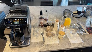 En køkkenbordplade med kaffemaskine, diverse flasker, glas og et spiseområde i baggrunden.