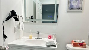 Een badkamer met een witte wastafel, een spiegel, een haardroger, een fles handzeep, een doos zakdoekjes en een toiletrolhouder.