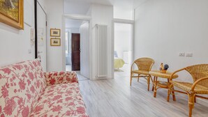 Ein Zimmer mit einem blumengemusterten Sofa, Korbstühlen, einem Heizkörper und einer Tür zu einem anderen Raum.