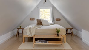 A bedroom with a sloped ceiling, a bed with pillows, a bedside table with a plant, and a window with curtains.