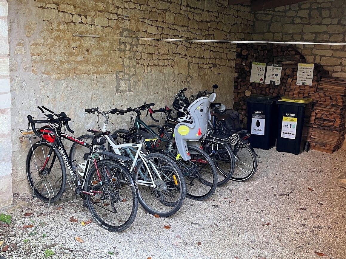 Fietsen staan tegen een stenen muur in een overdekte ruimte, met recyclingbakken en een stapel hout.