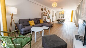 Una sala de estar moderna con un sofá gris, dos mesitas blancas, un sillón verde de mimbre y un televisor de pantalla plana.