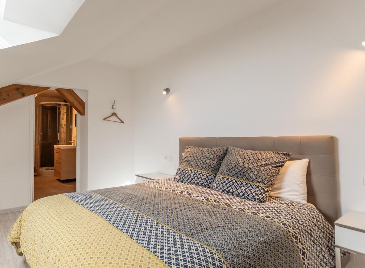 A bedroom with a bed, a bedside table, a hanging light, and a door leading to another room.