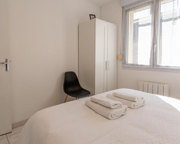 A bedroom with a bed, a black chair, a white wardrobe, and a window with blinds. - L'appart' de la Goutte D'or (Avize)