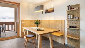 Une table à manger en bois avec des chaises, un luminaire suspendu, une étagère fixée au mur avec des bouteilles, et une vue à travers une porte vitrée.