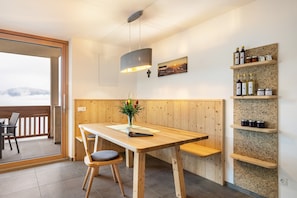 Une table à manger en bois avec des chaises, un luminaire suspendu, une étagère fixée au mur avec des bouteilles, et une vue à travers une porte vitrée.