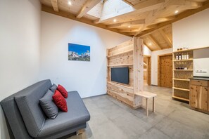 A modern living room with a grey sofa, red cushions, a flat-screen TV, wooden paneling, a bench, and a kitchen area with shelves and cabinets.